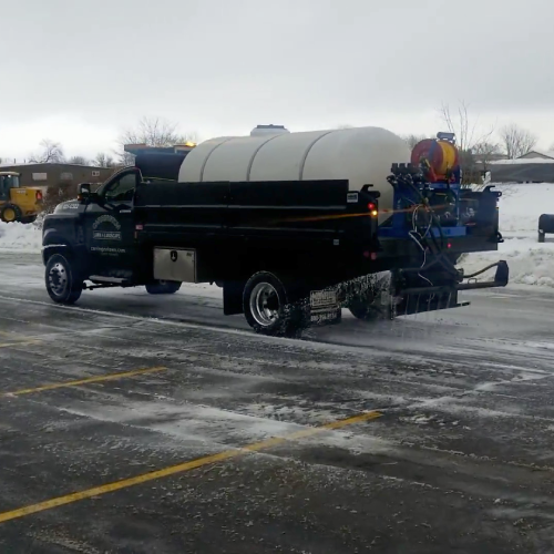 Medium-duty truck with a large brine tank sprays anti-ice across parking lanes during a commercial snow removal service by Carrington Lawn & Landscape in Middleton, Madison, and the greater Dane County area