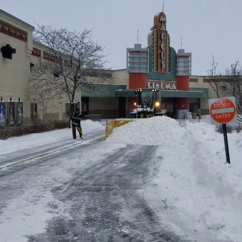 Loader with a box pusher clears snow at a cinema entrance, maintaining access lanes during a commercial snow removal service by Carrington Lawn & Landscape in Middleton, Madison, and the greater Dane County area