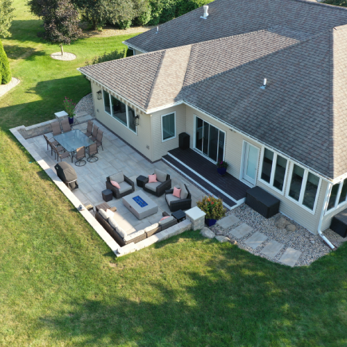 Oblique aerial of a completed linear paver patio with sectional lounge, gas fire table, dining area, seat walls, and garden steps—Waunakee outdoor living project by Carrington Lawn & Landscape in Madison metro area