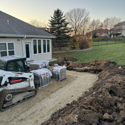 Construction scene with skid steer, compacted gravel base, open utility trench, and pallets of pavers staged beside the home—Waunakee outdoor living project by Carrington Lawn & Landscape in Madison metro area