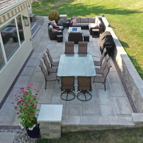 Close aerial view down the linear patio showing an eight-seat dining table, sectional lounge with fire table, grill area, and seat walls—Waunakee outdoor living project by Carrington Lawn & Landscape in Madison metro area