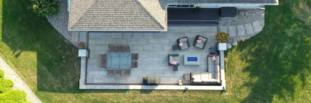 Overhead aerial showing full linear patio layout with dining zone, lounge around a rectangular fire table, grill position, and step access—Waunakee outdoor living project by Carrington Lawn & Landscape in Madison metro area