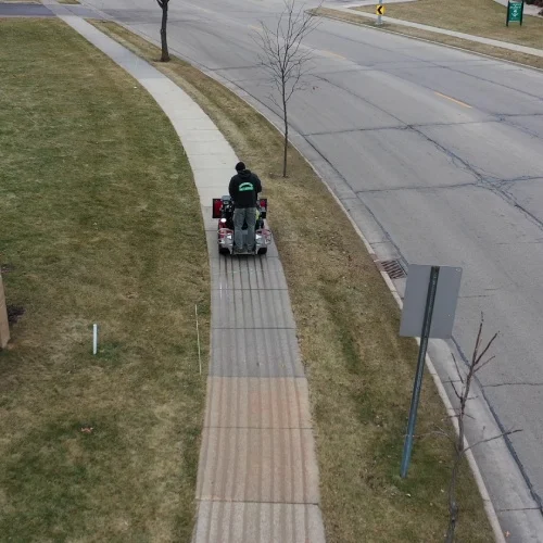 Operator uses a compact sidewalk machine to clear and deice a long walkway for pedestrian safety, certified snow professional by Carrington Lawn & Landscape