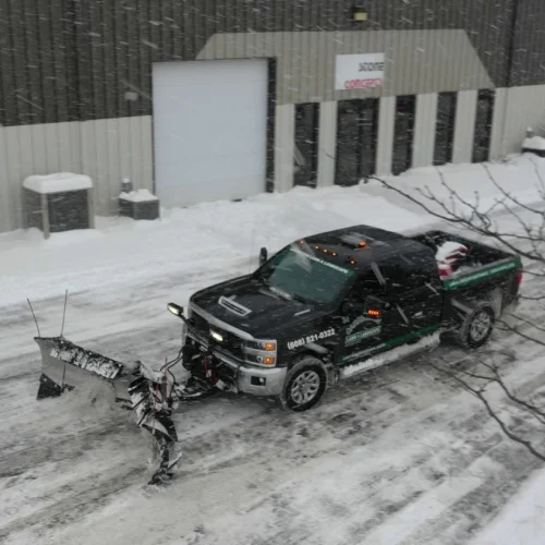 Pickup with front‑mounted plow clears a commercial drive during a snowstorm as part of snow and ice removal technology by Carrington Lawn & Landscape