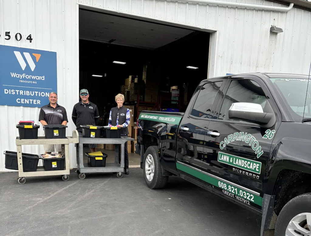 Carrington Lawn & Landscape team member infront of a warehouse with a sign "wayforward school supply drive results" in middleton, WI