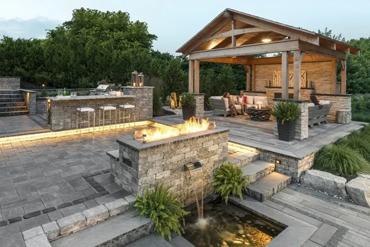 A backyard with a paved patio, a fire pit, a pergola, an outdoor kitchen, and a water feature with an elegant landscape lighting design in Middleton, WI