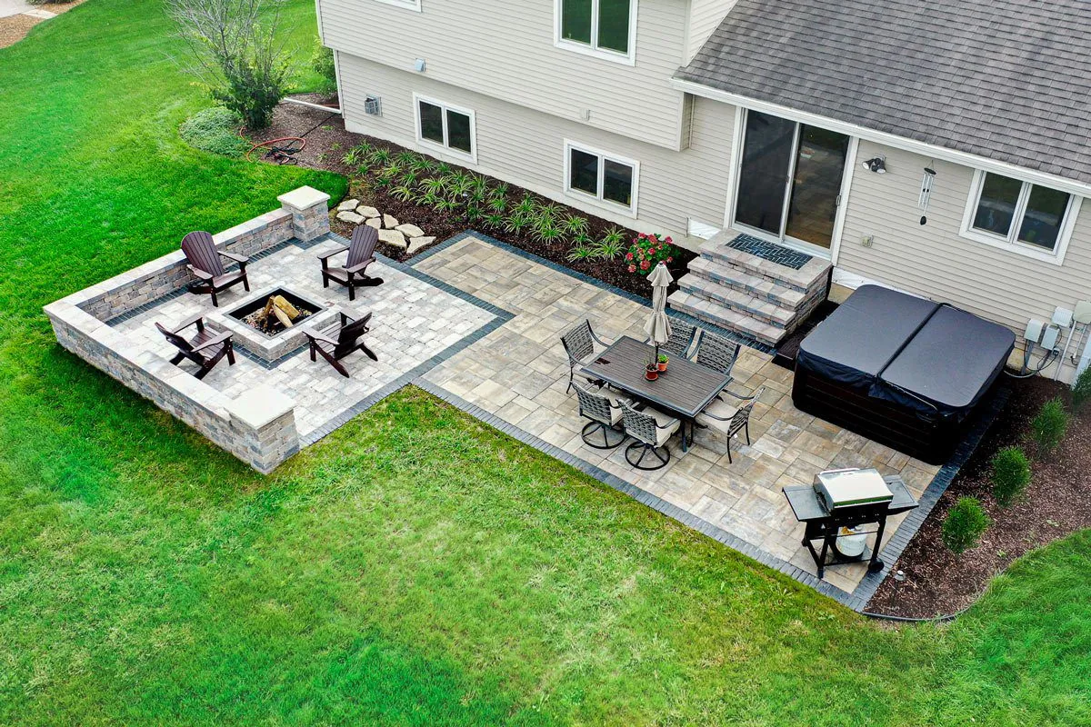Aerial view of a backyard featuring a two-tiered paver patio with a built-in fire pit, dining set, hot tub, and softscape landscaping in Middleton, WI