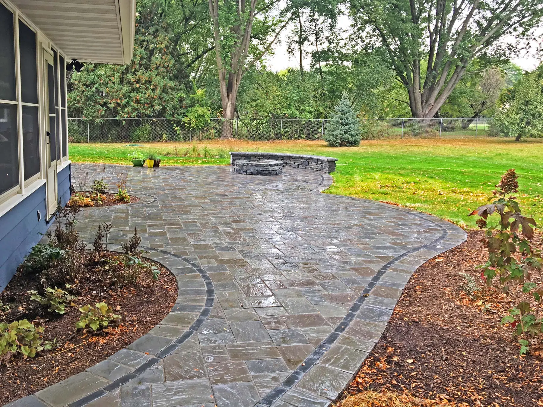 A curved hardcsape design stone patio leads to a built-in fire pit and seating area in a backyard.