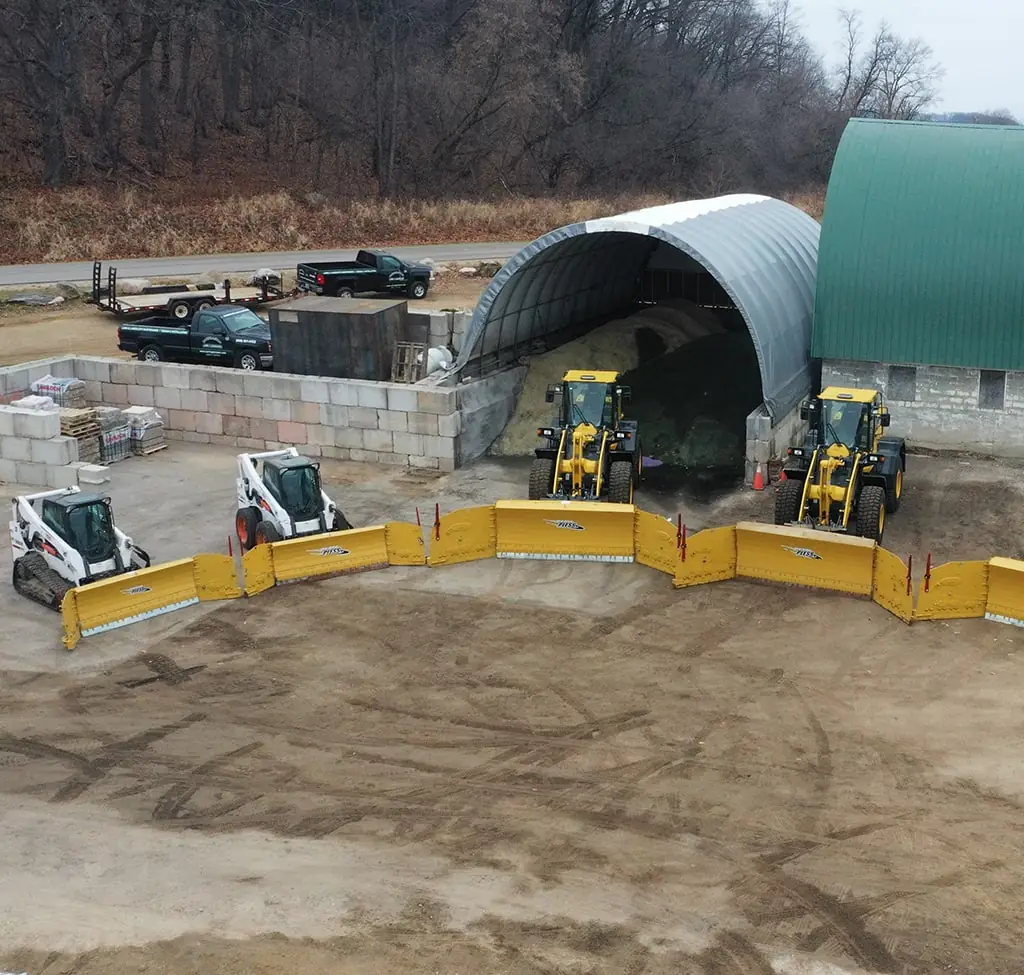 Aerial view of a construction site with three skid steer loaders and a larger loader, gravel piles, and a metal storage structure for snow and ice management in Middleton, WI