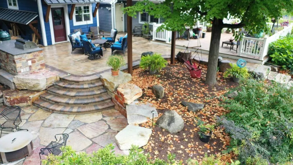 Overhead shot of a hardscape stone patio with blue outdoor furniture, a tiered stone staircase, and a landscaped area with a tree.