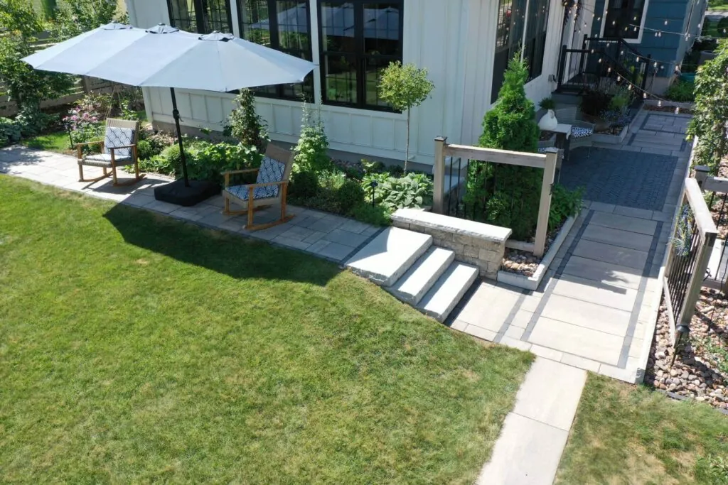 A hardscape design modern patio with seating, umbrella, and landscaping next to a white house in Middleton, WI