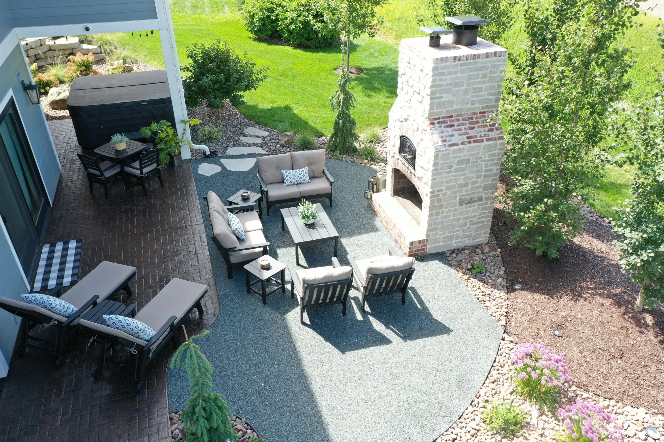 An aerial patio view of a large stone outdoor brick fireplace, comfortable cushioned seating on a gravel bed, and an adjacent brick paver patio with a hot tub showcasing a premium fire pit installation, completed as a professional fire pit installation in Middleton, Wisconsin by Carrington Lawn & Landscape.