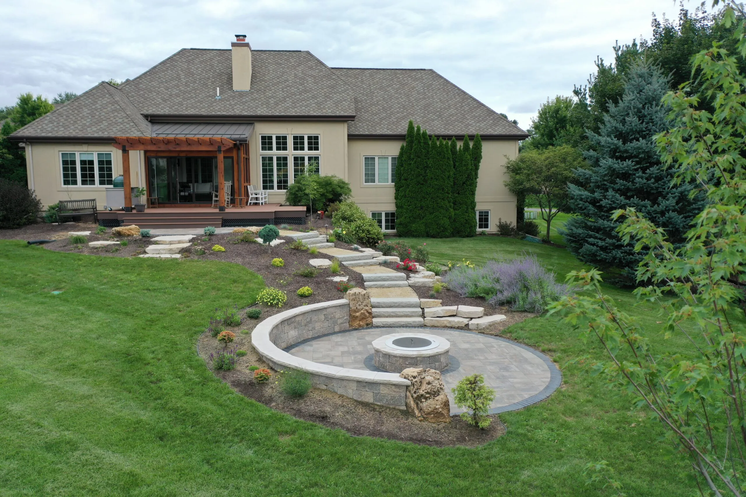 A sloped backyard featuring a custom circular paver patio with a stone fire pit, a curved retaining wall, and natural stone steps showcasing elegant fire pit renovation ideas, completed as a professional fire pit renovation in Middleton, Wisconsin by Carrington Lawn & Landscape.