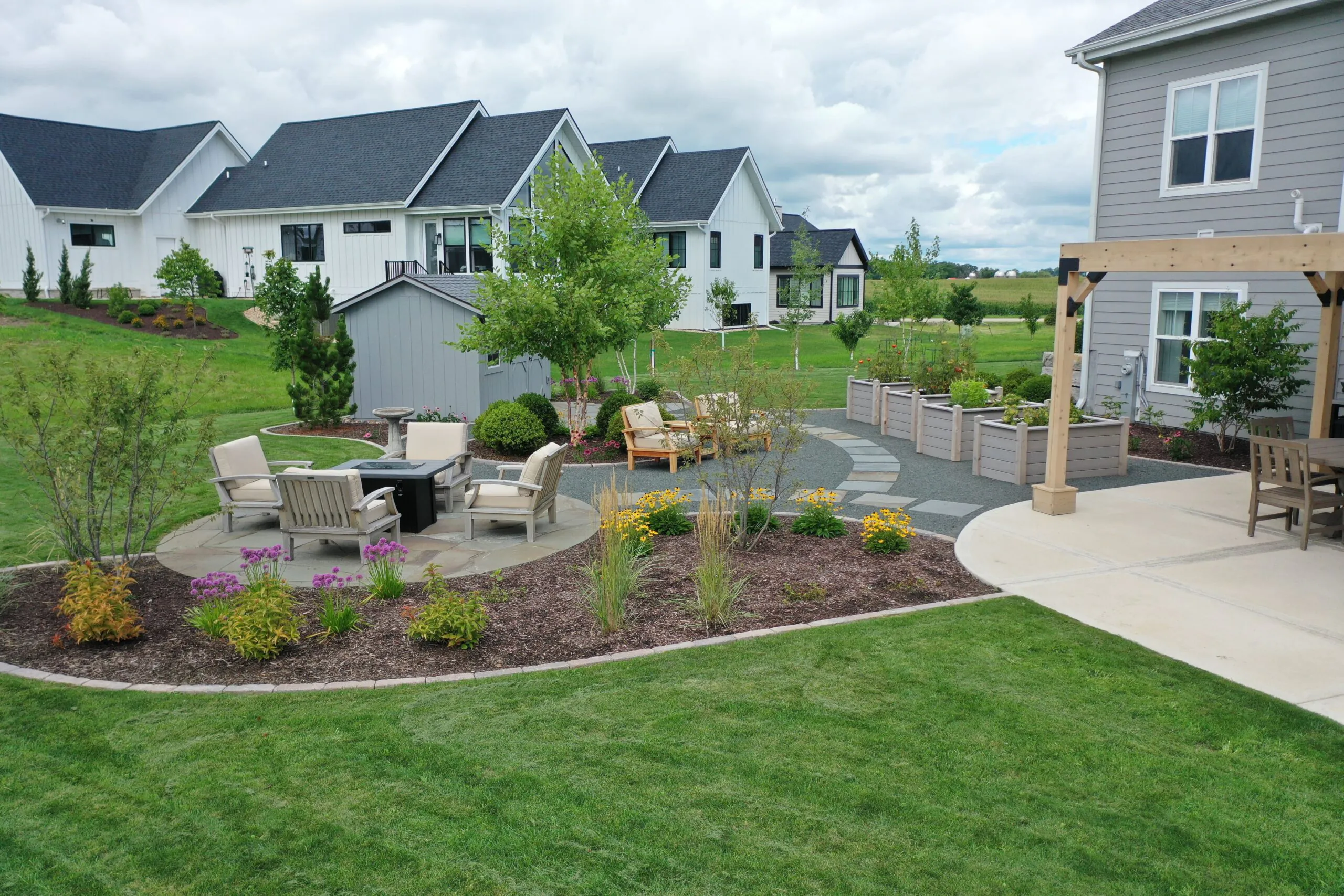 A wide elevated view of a fully landscaped suburban backyard with a flagstone patio, fire pit seating area, and mulched flower beds featuring a custom softscape landscape design in Westport WI, featuring custom softscape landscape design in Middleton, Wisconsin by Carrington Lawn & Landscape.