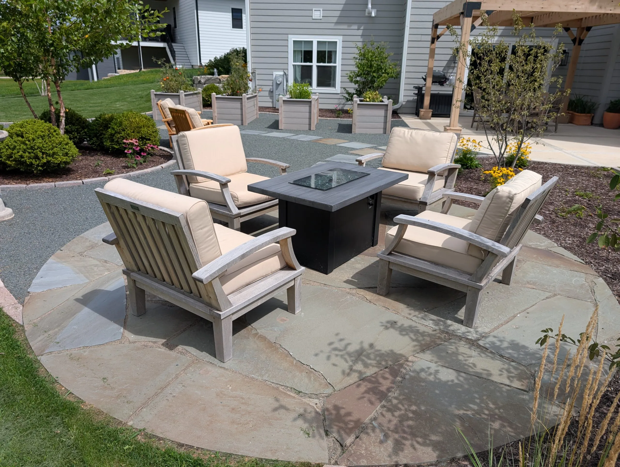 A close-up view of a circular flagstone patio featuring four wooden armchairs with cream cushions surrounding a dark rectangular fire pit table, integrated into a custom softscape landscape design in Westport WI, featuring custom softscape landscape design in Middleton, Wisconsin by Carrington Lawn & Landscape.