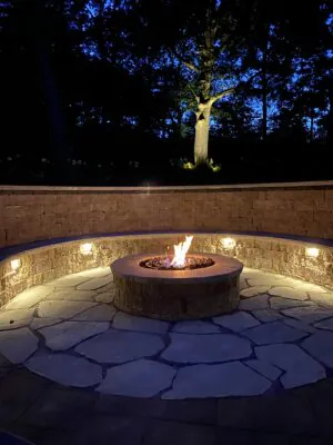 A glowing stone fire pit lit up at night on a flagstone patio with an integrated curved stone seating wall and custom landscape lighting highlighting inspiring fire pit renovation ideas, completed as a professional fire pit renovation in Middleton, Wisconsin by Carrington Lawn & Landscape.