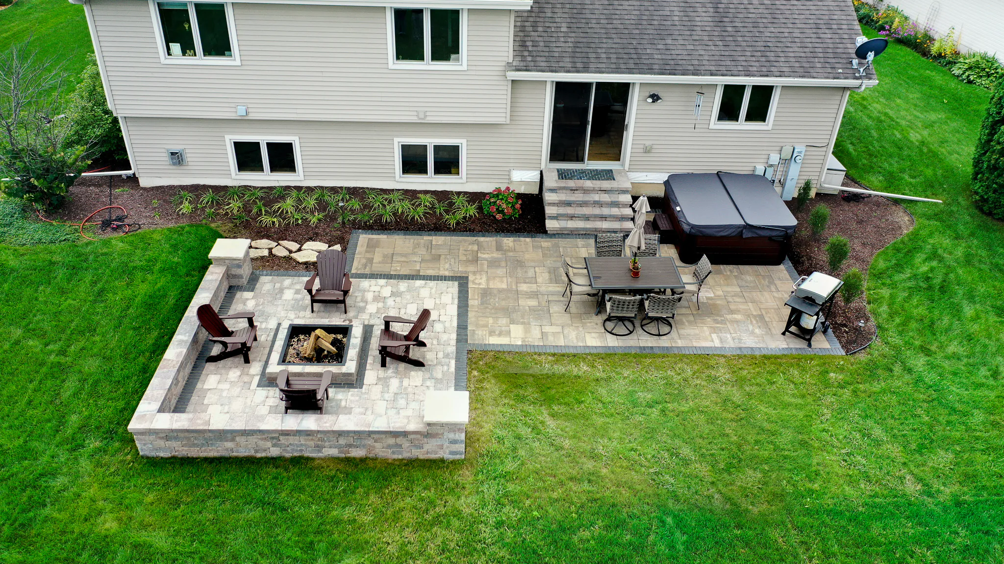 An aerial view of a custom multi-level stone paver patio featuring a raised square stone fire pit area with dark brown Adirondack chairs, an outdoor dining set, and a hot tub, completed as a professional fire pit installation in Middleton, Wisconsin by Carrington Lawn & Landscape