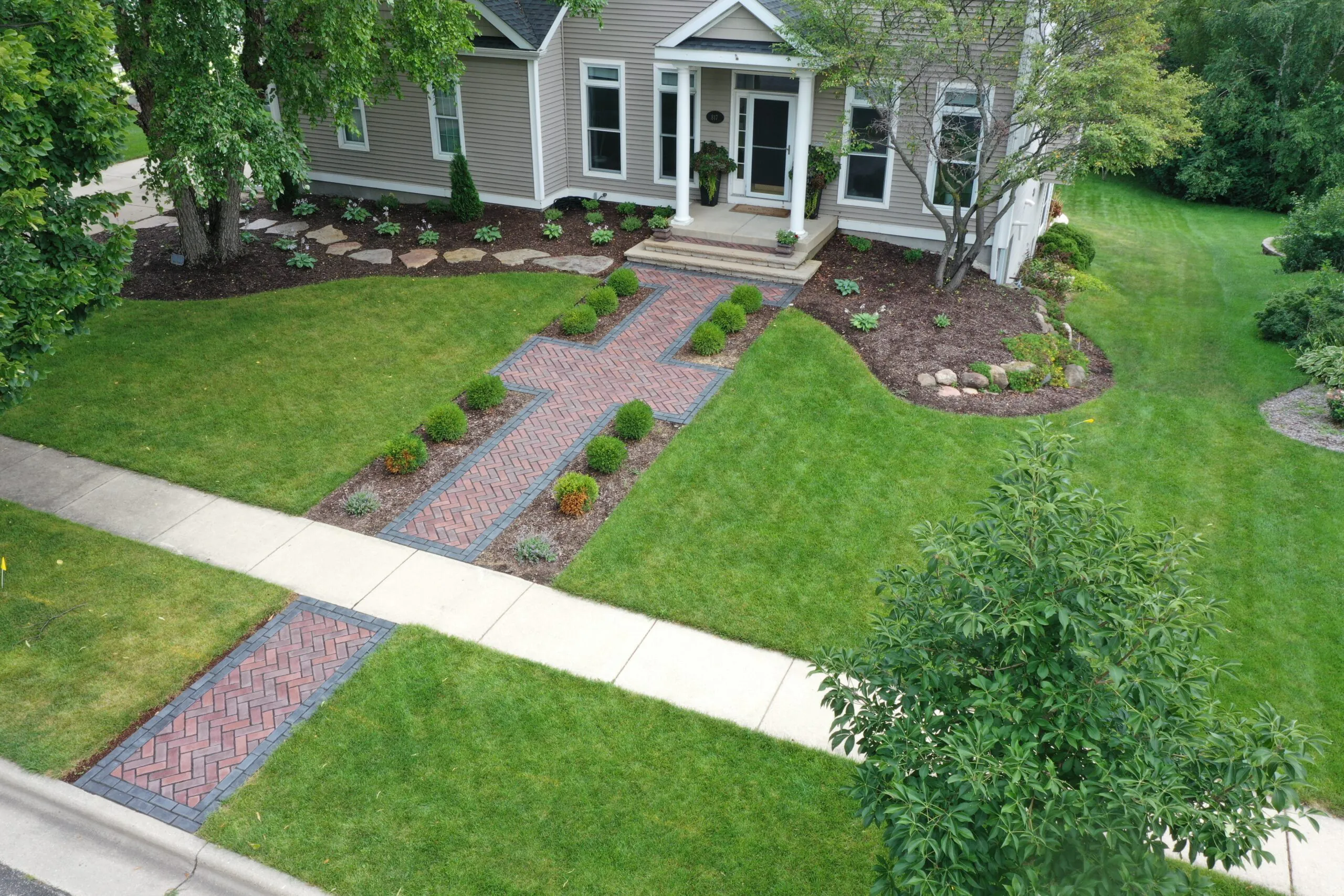 An aerial drone view of a red herringbone paver walkway connecting the curb and front porch through a lush lawn, highlighting material choices in concrete vs paver patio and walkway design, completed as a custom paver installation in Middleton, Wisconsin by Carrington Lawn & Landscape.