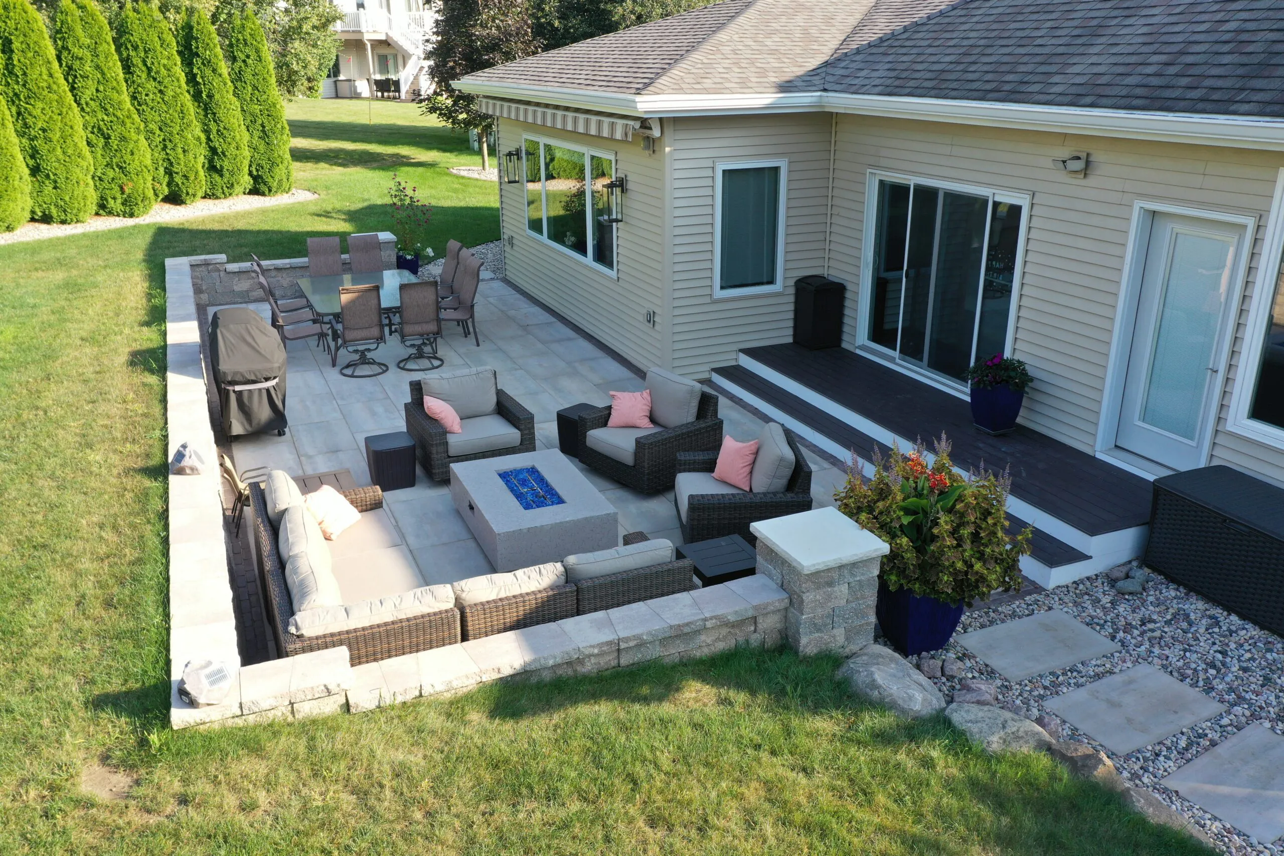 An aerial view of a large stone paver patio featuring a modern rectangular gas fire pit table with blue glass rock media, outdoor sectional seating, and a dining area highlighting gas fire pit vs wood burning designs, built as a fire pit installation in Middleton, Wisconsin by Carrington Lawn & Landscape.