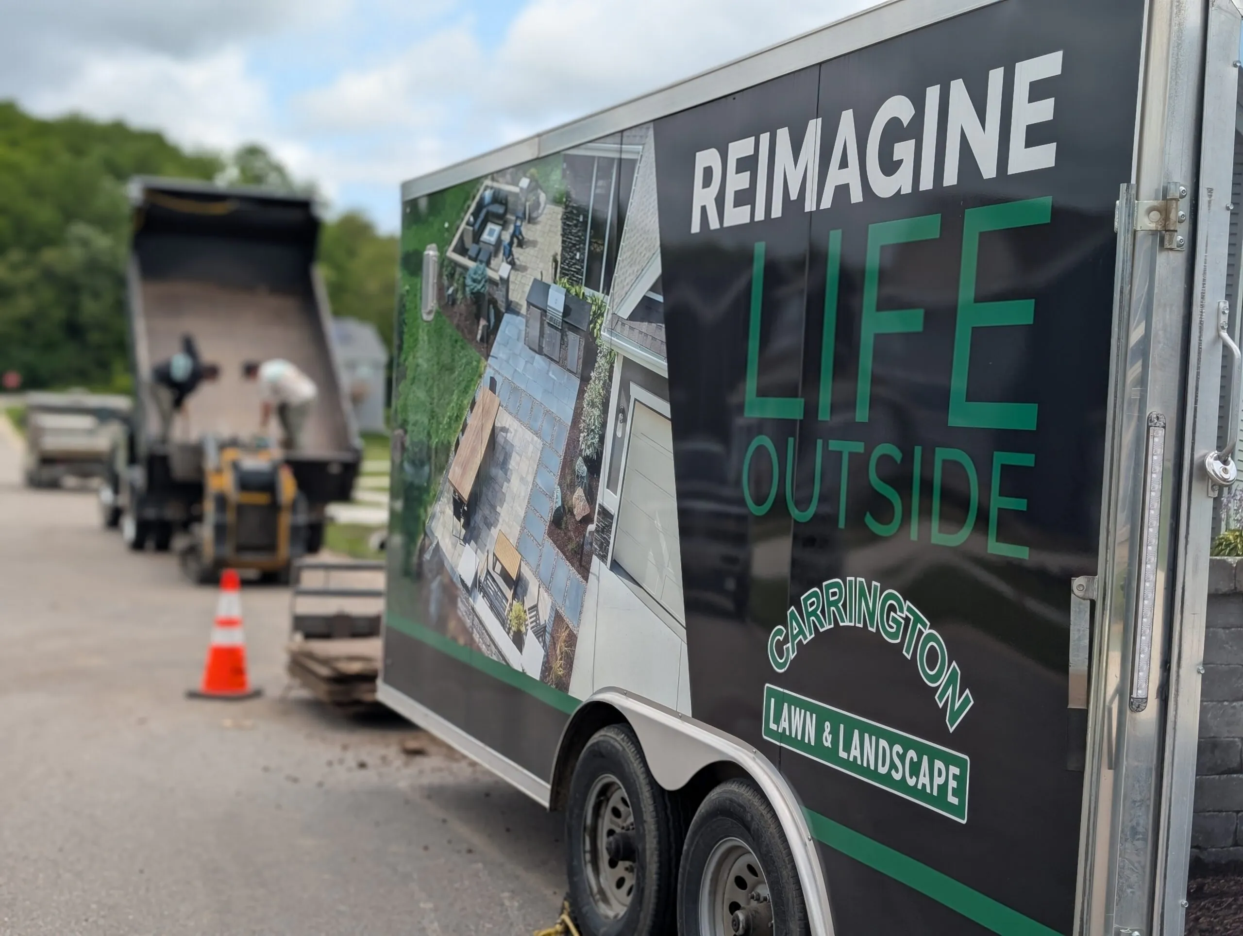 A branded work trailer parked on a street near a dump truck and crew working on-site during key landscape design process steps, managed as part of the landscape design process in Middleton, Wisconsin by Carrington Lawn & Landscape.