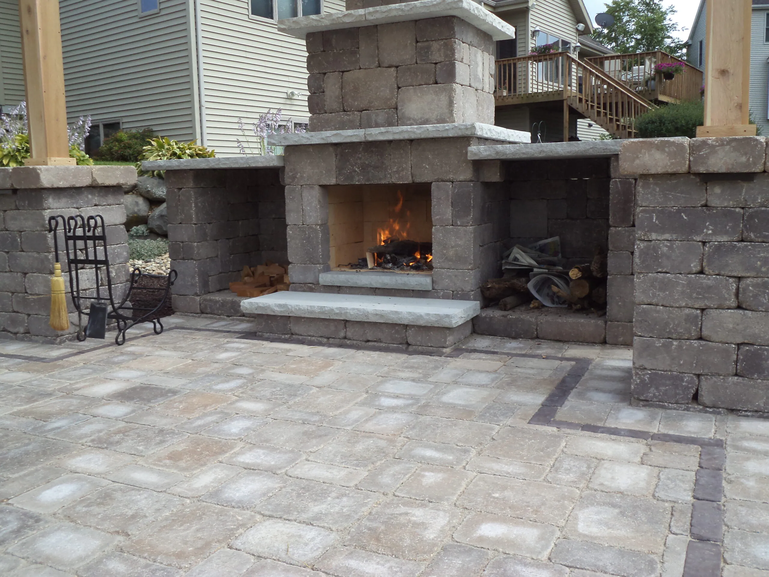 A custom stone outdoor fireplace with a wood-burning fire, built-in firewood storage nooks, and a paver patio demonstrating gas fire pit vs wood burning options, built as a fire pit installation in Middleton, Wisconsin by Carrington Lawn & Landscape.