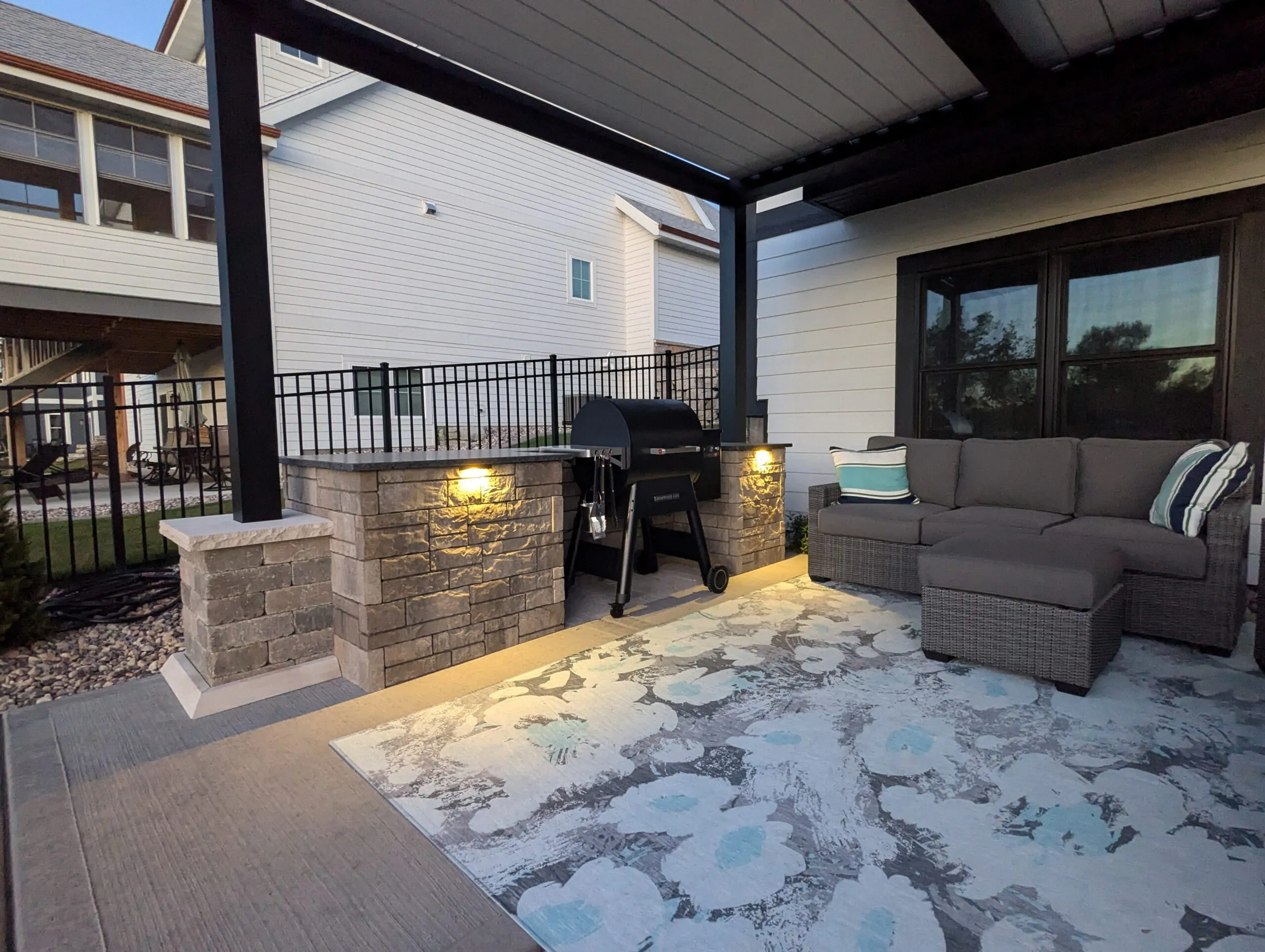 A covered outdoor patio and outdoor kitchen area with a smoker grill, gray sectional sofa, and warm stone pillar task lights displaying practical landscape lighting ideas, installed as professional landscape lighting in Middleton, Wisconsin by Carrington Lawn & Landscape.